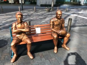 Two golden statues of smiling rugby players sit on a bench.
