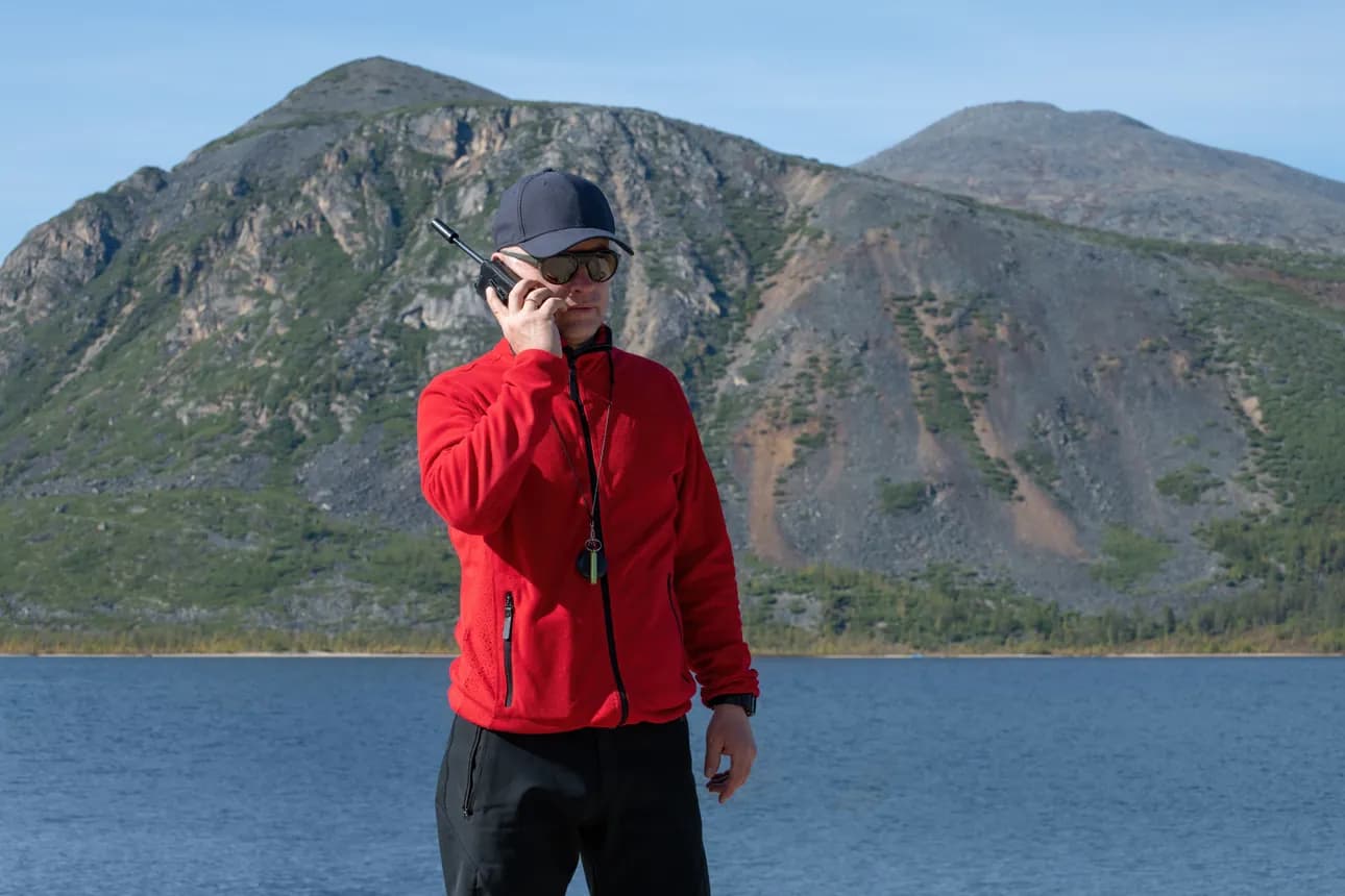 A man in a red fleece uses a handheld satellite phone beside a lake in remote mountain terrain.