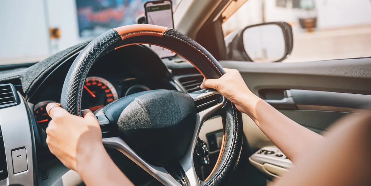 A driver's hands on a car steering wheel, with a phone mounted on the windscreen ahead.